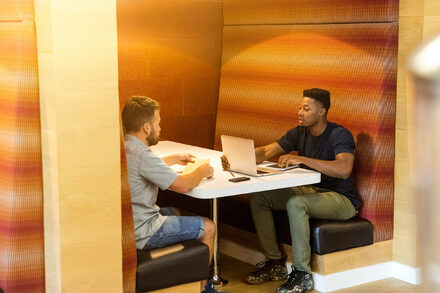 Two young men sat in a cafe booth engaged in conversation while looking at a laptop screen